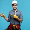 Maintenance, repair works and renovation concept. Stylish young European builder in gloves and safety helmet working on construction site, posing at blue wall and pointing index fingers away